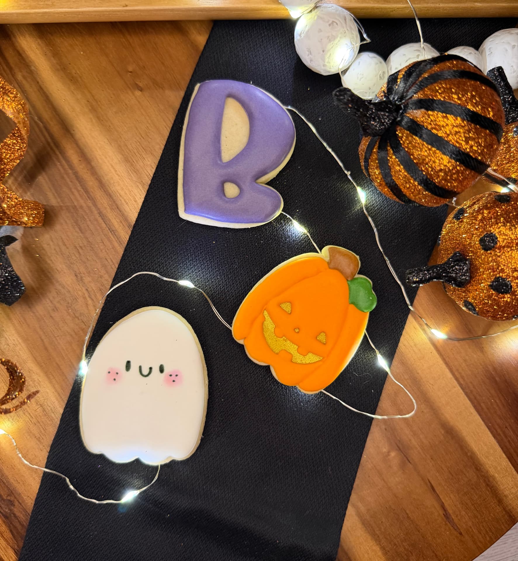 Halloween sugar cookies — a ghost, a jack-o'-lantern, and a 'Boo' letter on a black runner with fairy lights